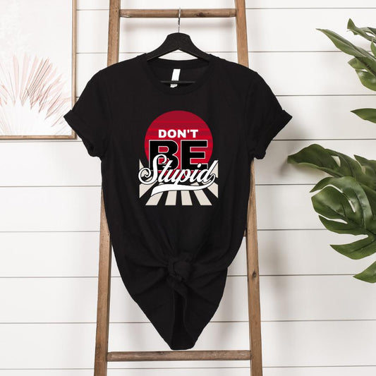 Black "Don't Be Stupid" t-shirt with red and white graphic design hanging on a wooden ladder against a white shiplap wall with plants nearby