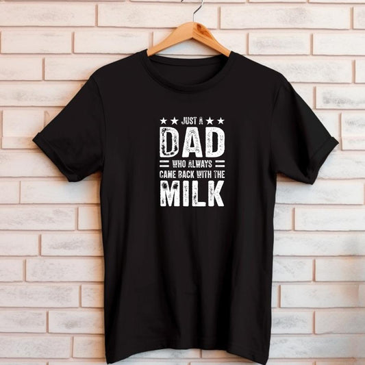 Black t-shirt with white text saying "Just a Dad who always came back with the milk" hanging on wooden hanger against brick wall background