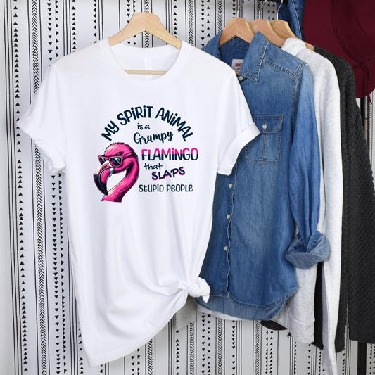 White t-shirt with humorous "My Spirit Animal Is a Grumpy Flamingo" design featuring a pink flamingo illustration, displayed on a hanger with casual clothes behind.