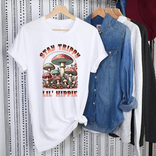 White Stay Trippy Lil' Hippie T-shirt with colorful psychedelic mushroom graphic hanging next to denim and casual shirts.