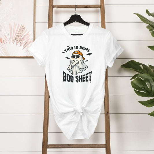 White This Is Some Boo Sheet T-Shirt with a cartoon ghost wearing sunglasses and a bandana printed on the front, displayed on a wooden ladder against a white shiplap wall with plants and artwork nearby