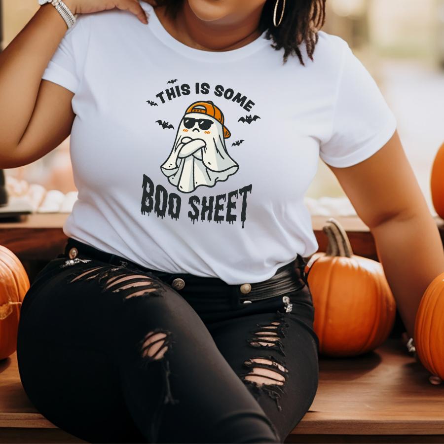 Woman wearing a white "This Is Some Boo Sheet" ghost pun t-shirt with sunglasses and a hat, sitting among pumpkins